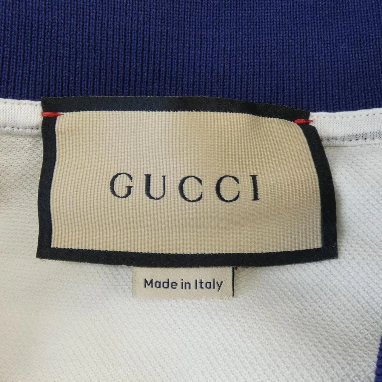 Beige Gucci neck label with a black border and red accents at the top corners, GUCCI in black serif capitals, above a separate cream tab reading Made in Italy