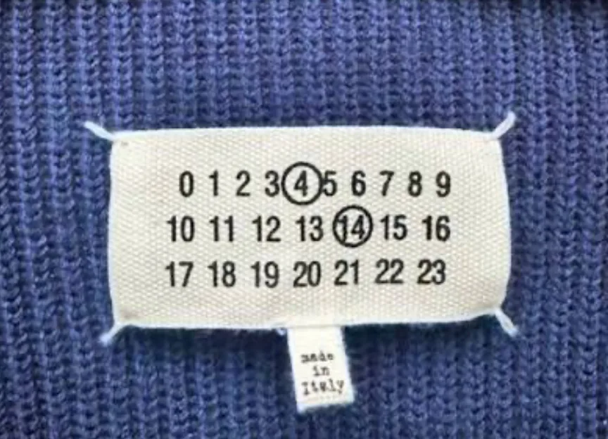 Maison Margiela numbers label on a blue knit, showing 0 to 23 in three rows with 4 and 14 circled, tacked at four corners with white thread, above a small tab reading made in Italy