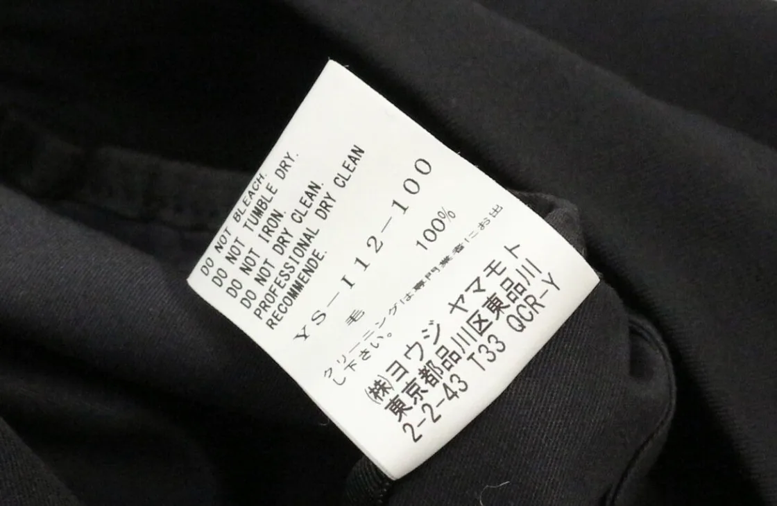 Yohji Yamamoto care label printed sideways, reading YS-I12-100, 毛 100%, and English instructions DO NOT BLEACH, DO NOT TUMBLE DRY, DO NOT IRON, DO NOT DRY CLEAN, PROFESSIONAL DRY CLEAN RECOMMENDED, with the company name and Shinagawa address in Japanese